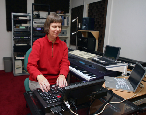 Picture: Veronica in her studio! She is sitting with her hands on the controls of the Radar hard disk recorder; appears to be listening, with a half-smile; surrounded by computer and synthesizer keyboards. In the background you see speakers and other gear.